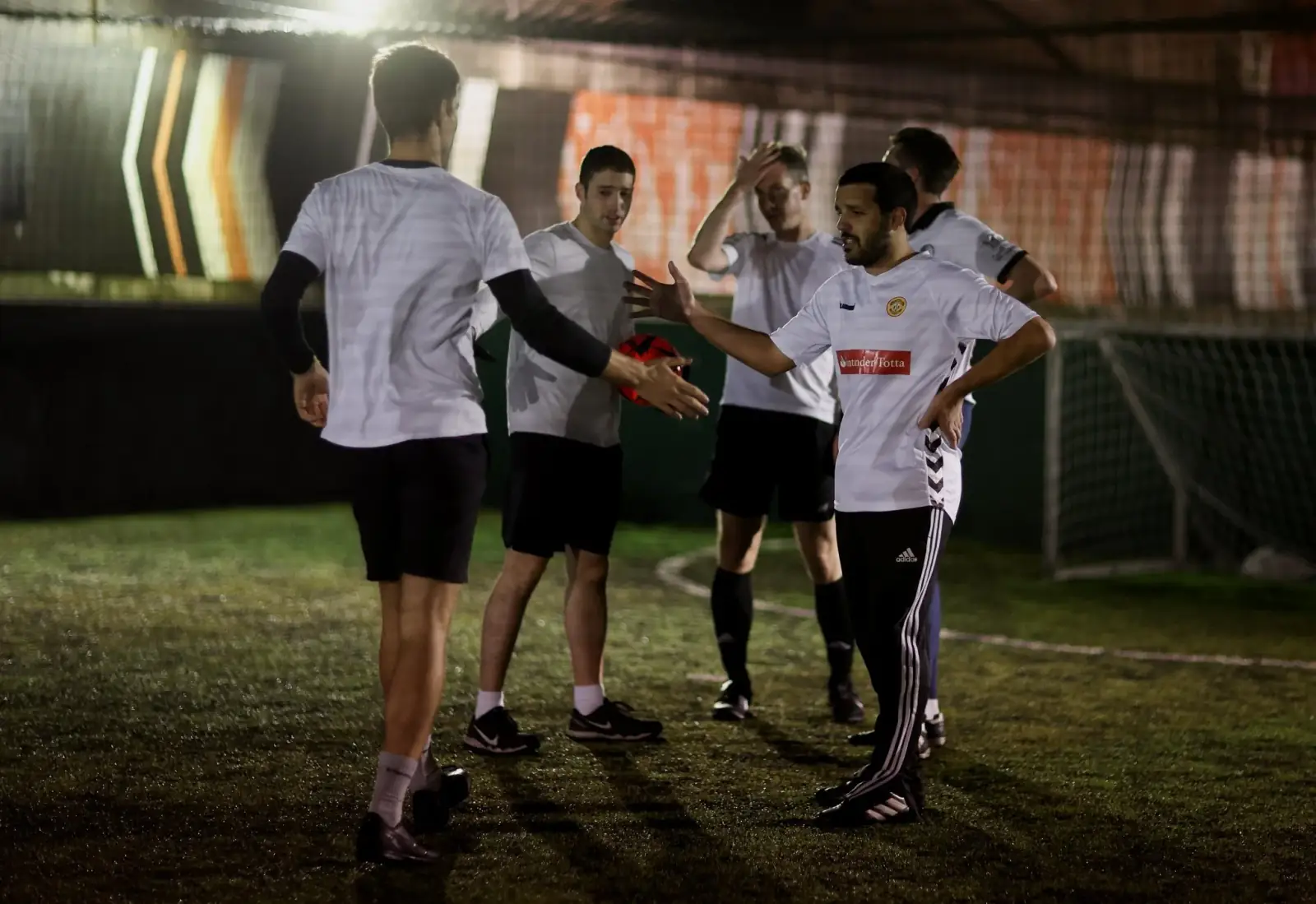 A group of players in white shirts gathering after a match, one reaching out to shake hands beside an Adidas-branded pitch wall.