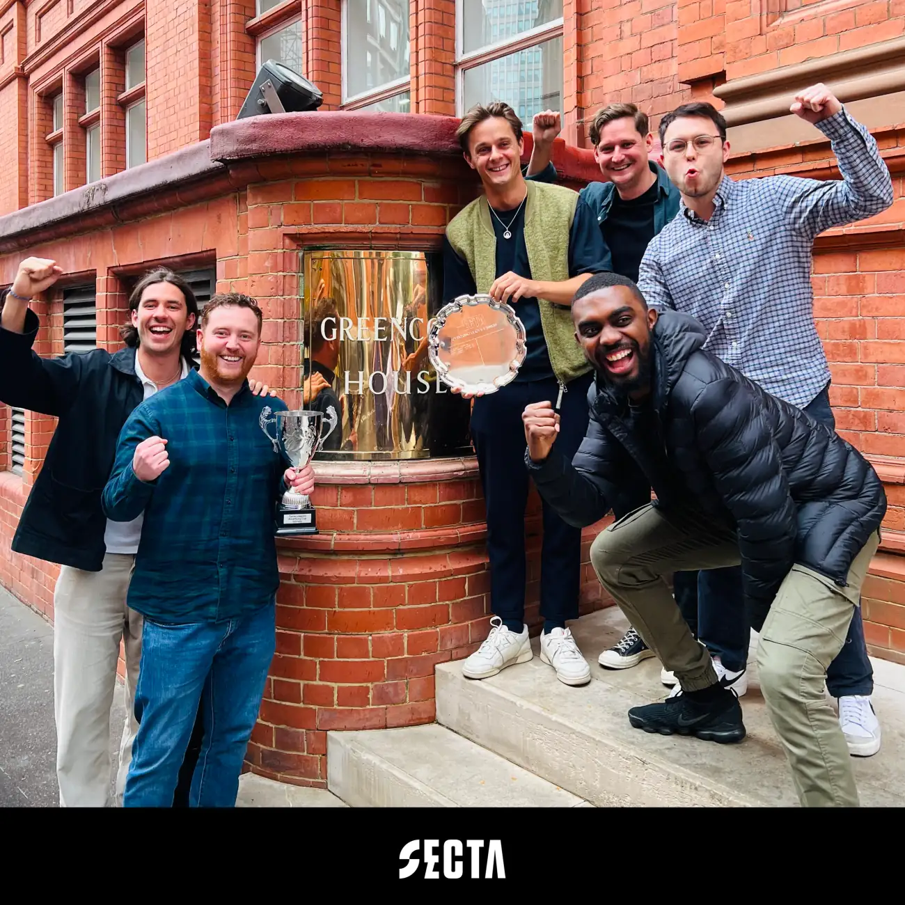 Six winning teammates cheering outside a red-brick London office building, one holding a silver trophy and another a runners-up salver.