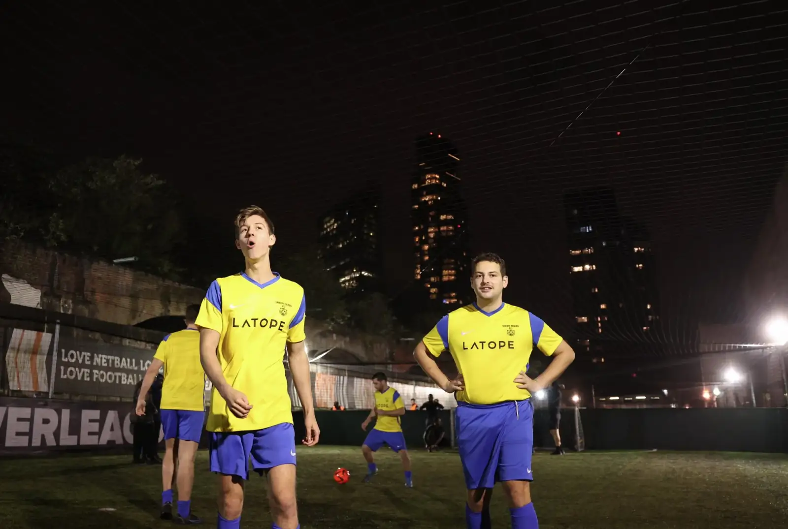 A women's five-a-side team gathered pitchside under a railway arch at night, London tower blocks lit up in the background.