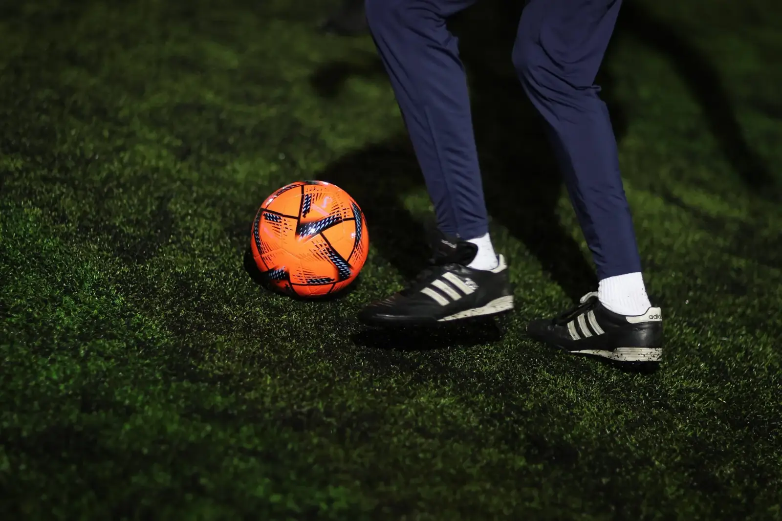 Close-up of a football and a player's boots mid-dribble on a floodlit artificial pitch.