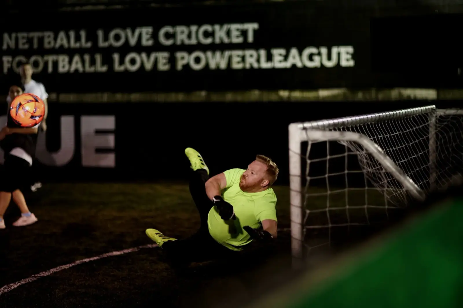 A goalkeeper diving full stretch to save a shot during a five-a-side match, the ball flying past the post.