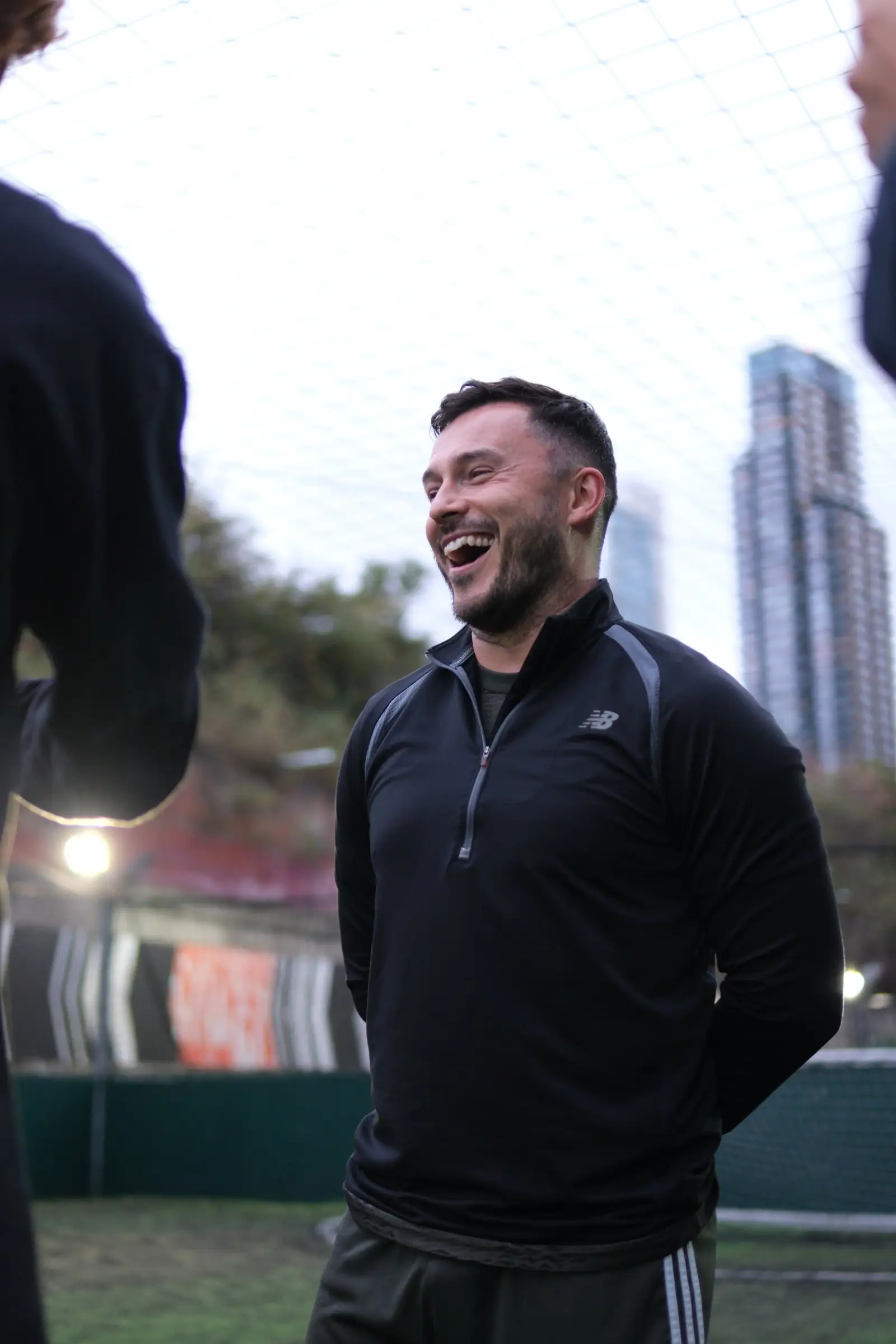 A player laughing mid-conversation pitchside at a five-a-side venue, London skyscrapers rising behind the goal netting.