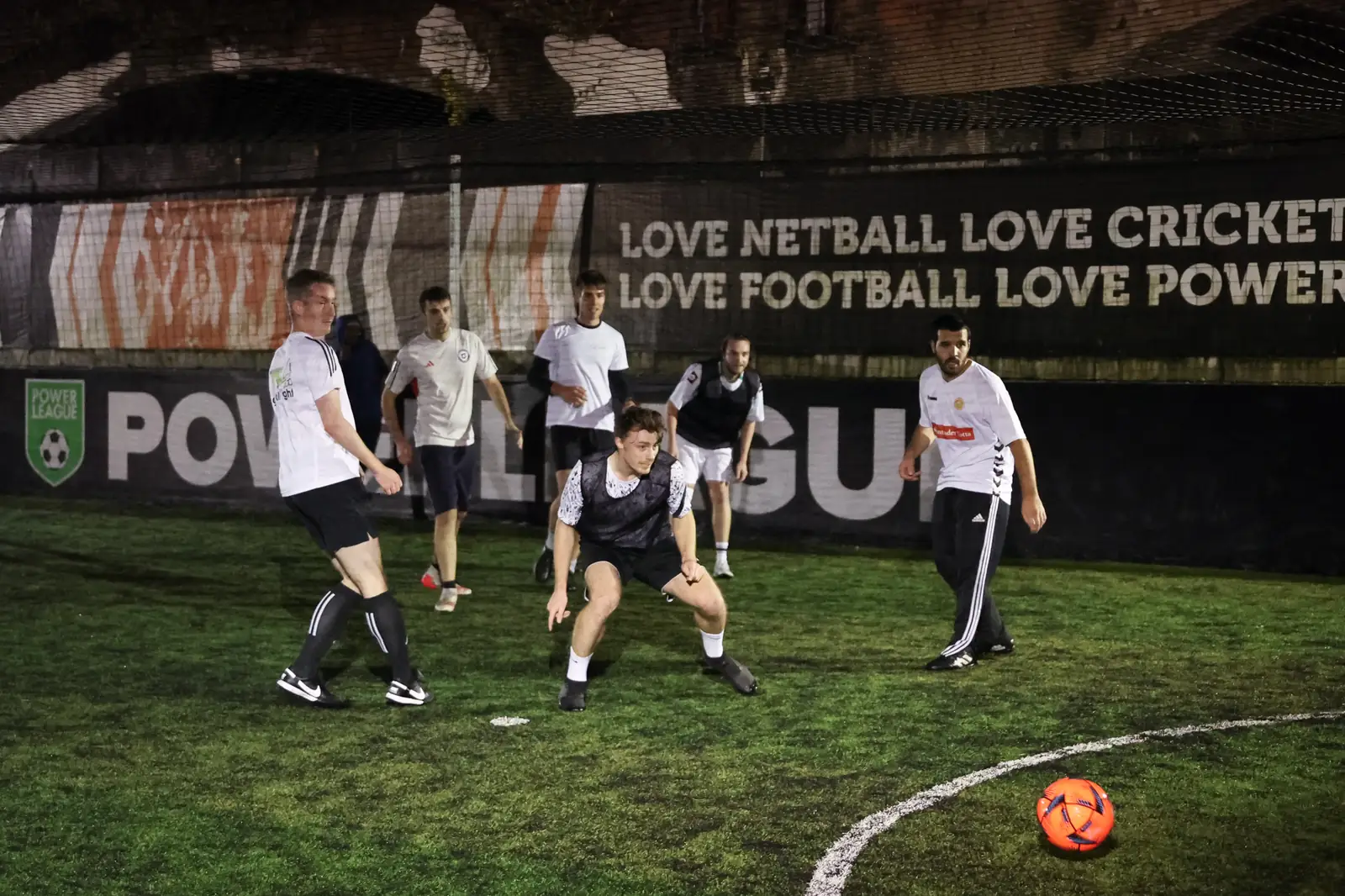 Players in white and black kit chasing a loose ball across a floodlit five-a-side pitch, Powerleague boards behind them.