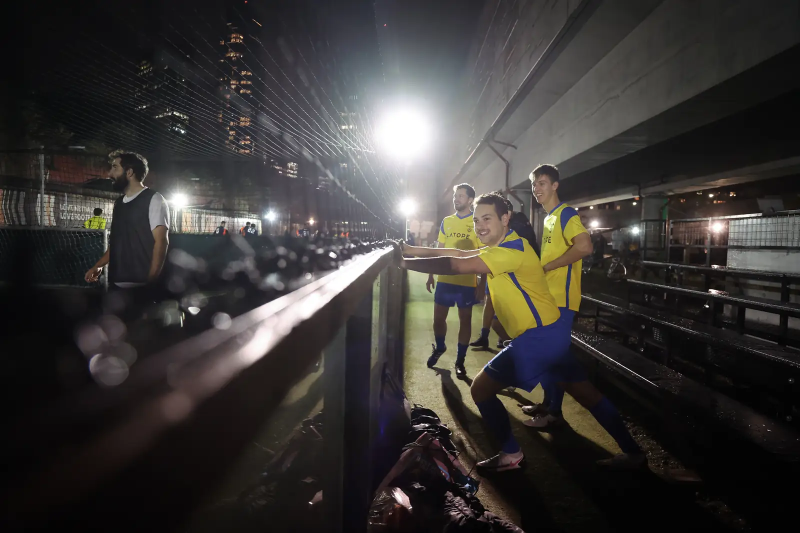 Players in yellow and blue kit cheering a teammate from the pitchside rail under bright floodlights, tower blocks behind.