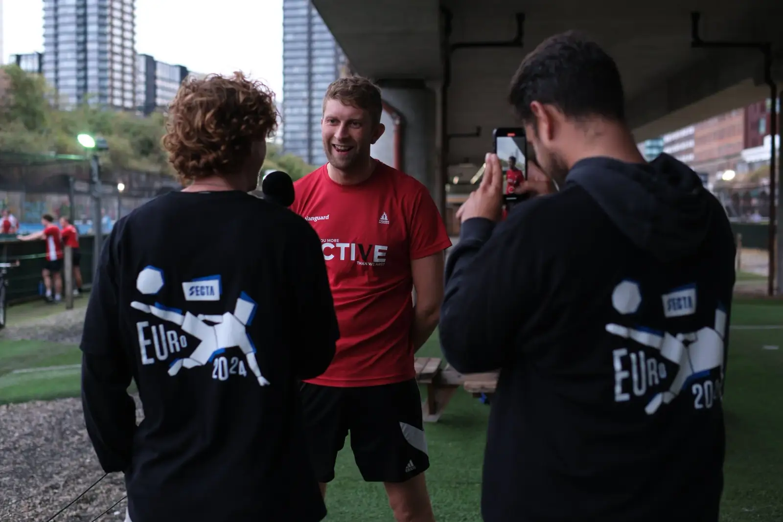 A content crew filming a smiling player being interviewed with a microphone after a match, a phone camera recording in the foreground.