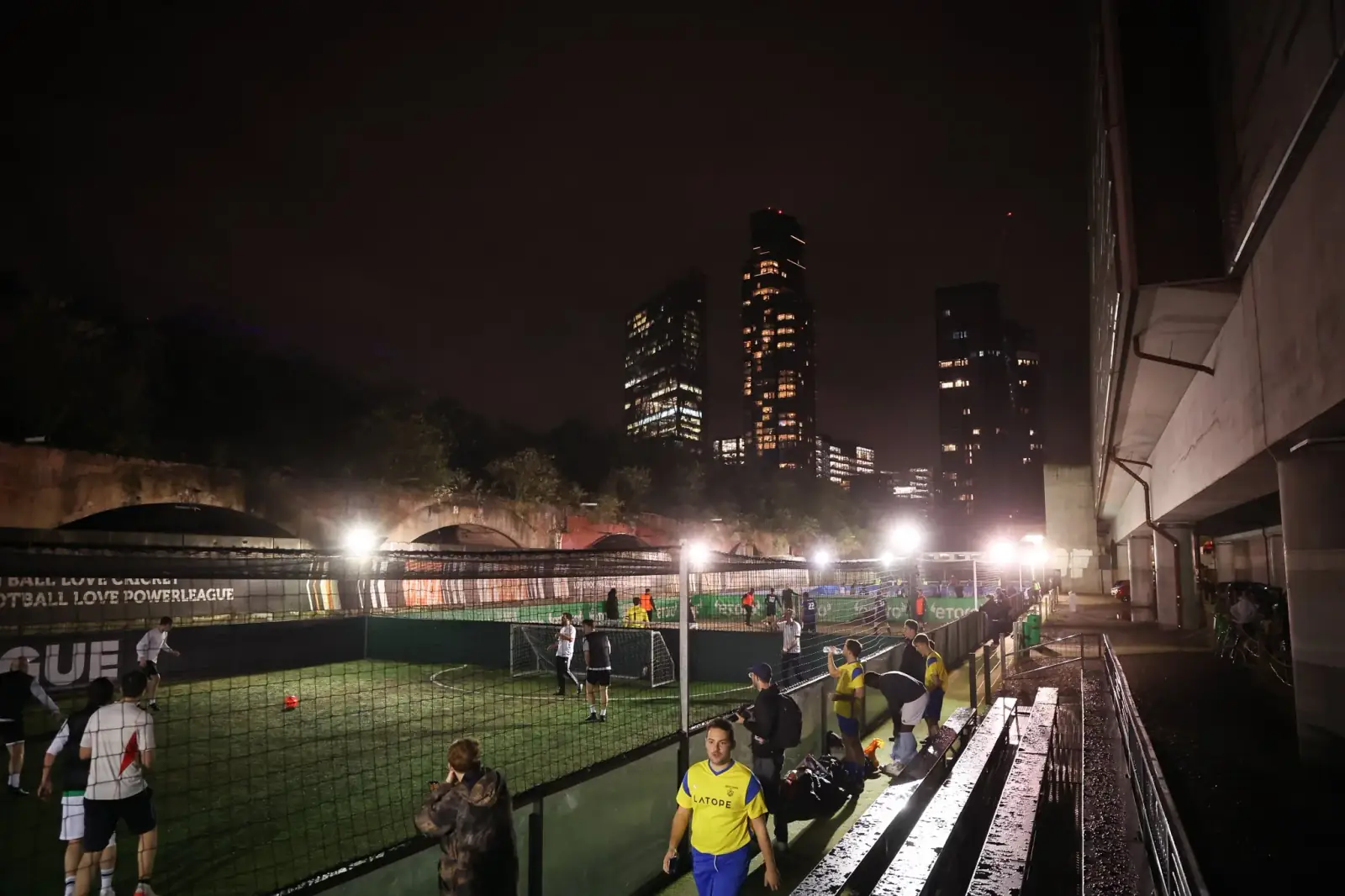 A wide view of a floodlit five-a-side venue at night with London tower blocks rising behind it and players gathering pitchside.