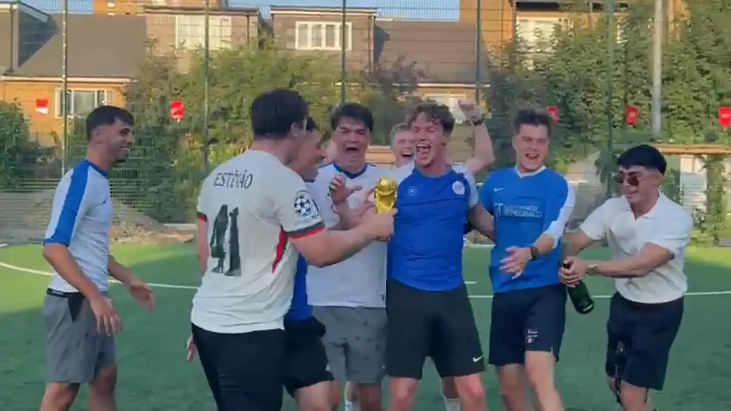 Gardiner & Theobald players in blue shirts crowding round a gold World Cup trophy on a five-a-side pitch, shouting and laughing, a champagne bottle held out at the right.