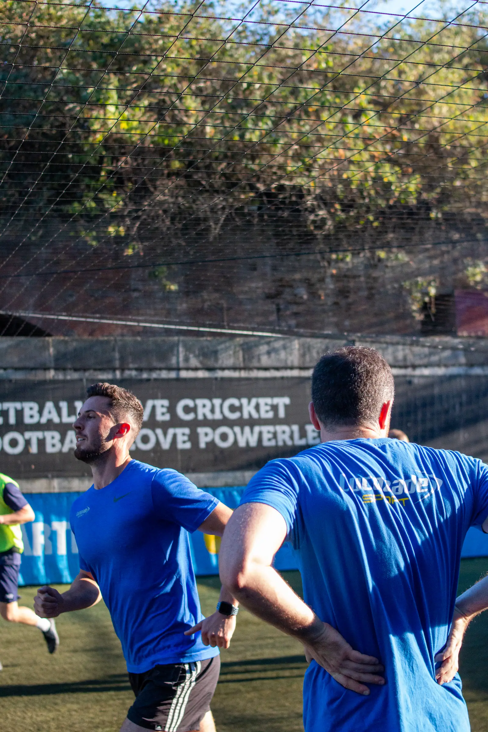 Players in blue kit competing during the Lucozade versus adam&eve match on a sunlit London pitch.