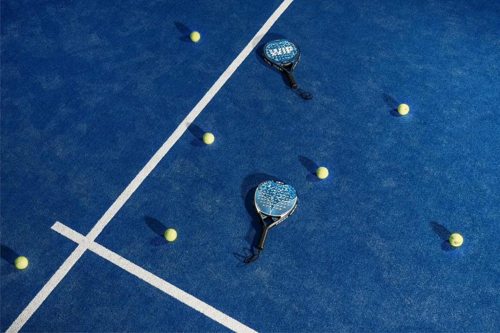 Overhead view of a blue padel court with two rackets and scattered balls on the surface.