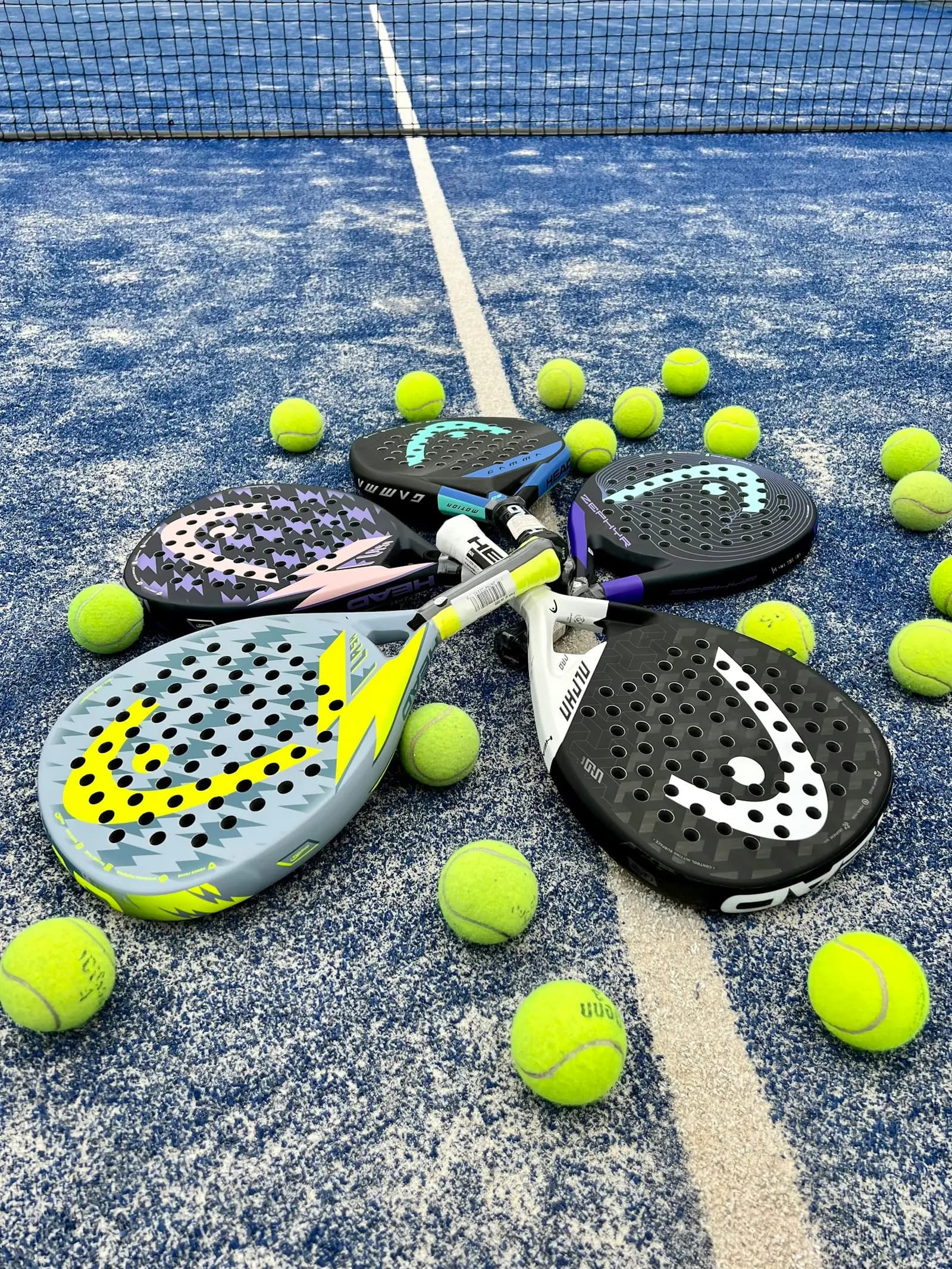 A pile of padel rackets and balls resting on a blue court beside the net.